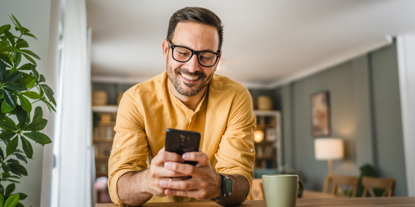Ein Mann in entspannter Atmosphäre zuhause nutzt sein Smartphone und freut sich über die Vorteile moderner Telekommunikationslösungen – zum Beispiel durch Zusatzoptionen wie die Mobil-Flat.