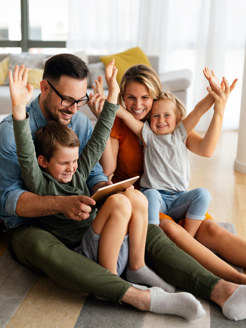 Familie sitzt gemeinsam im Wohnzimmer und nutzt ein Tablet, während Eltern und Kinder fröhlich die Hände heben.
