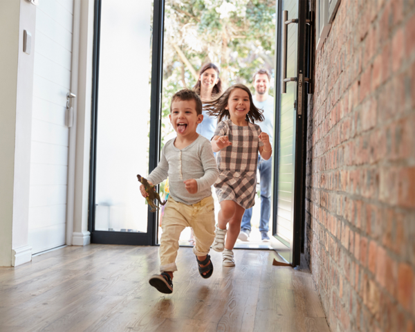 Zwei fröhliche Kinder rennen in ein neues Zuhause, gefolgt von ihren Eltern – Symbolbild für einen Familien-Neubau mit Glasfaseranschluss.
