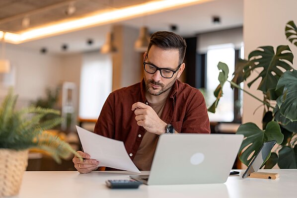 Ein Mann mit Brille arbeitet konzentriert am Schreibtisch. Er hält ein Dokument in der Hand, während ein Laptop vor ihm steht.