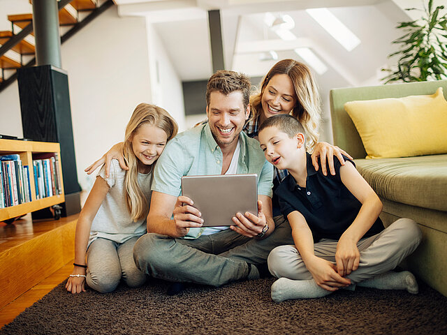 Eine Familie sitzt gemeinsam im Wohnzimmer auf dem Boden und schaut lächelnd auf ein Tablet.