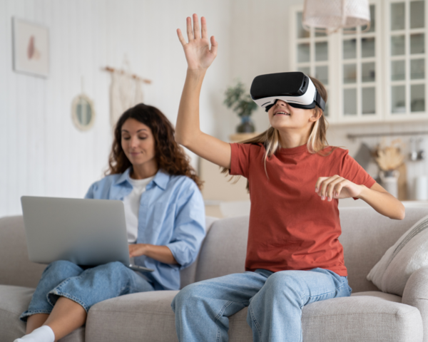 Frau mit Laptop auf dem Schoß und Mädchen mit VR Brille sitzen auf dem Sofa 