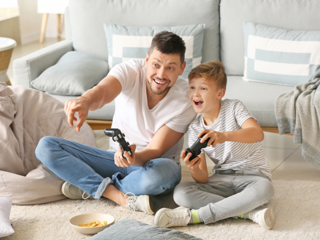 Vater und Sohn spielen gemeinsam begeistert mit Spielekonsole im Wohnzimmer.