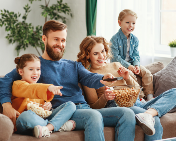 Vierköpfige Familie sitzt auf dem Sofa, der Vater hält eine Fernbedienung in der Hand