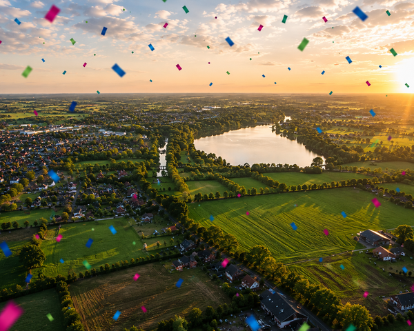 Eine Luftaufnahme zeigt eine ländliche Region mit einem See im Abendlicht, über der buntes Konfetti schwebt und eine festliche Stimmung vermittelt.