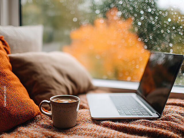 Ein Laptop und eine Tasse mit Getränk liegen auf einer Decke am Fenster, während draußen buntes Herbstlaub und Regentropfen zu sehen sind.