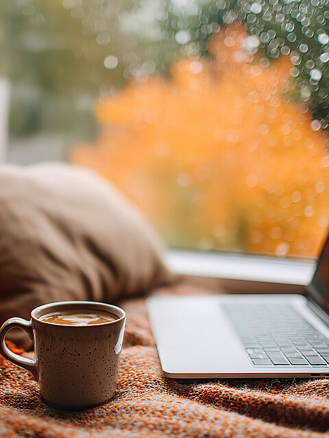Ein Laptop und eine Tasse mit Getränk liegen auf einer Decke am Fenster, während draußen buntes Herbstlaub und Regentropfen zu sehen sind.