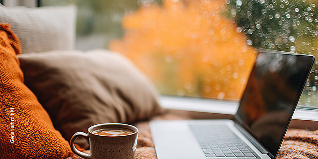 Ein Laptop und eine Tasse mit Getränk liegen auf einer Decke am Fenster, während draußen buntes Herbstlaub und Regentropfen zu sehen sind.
